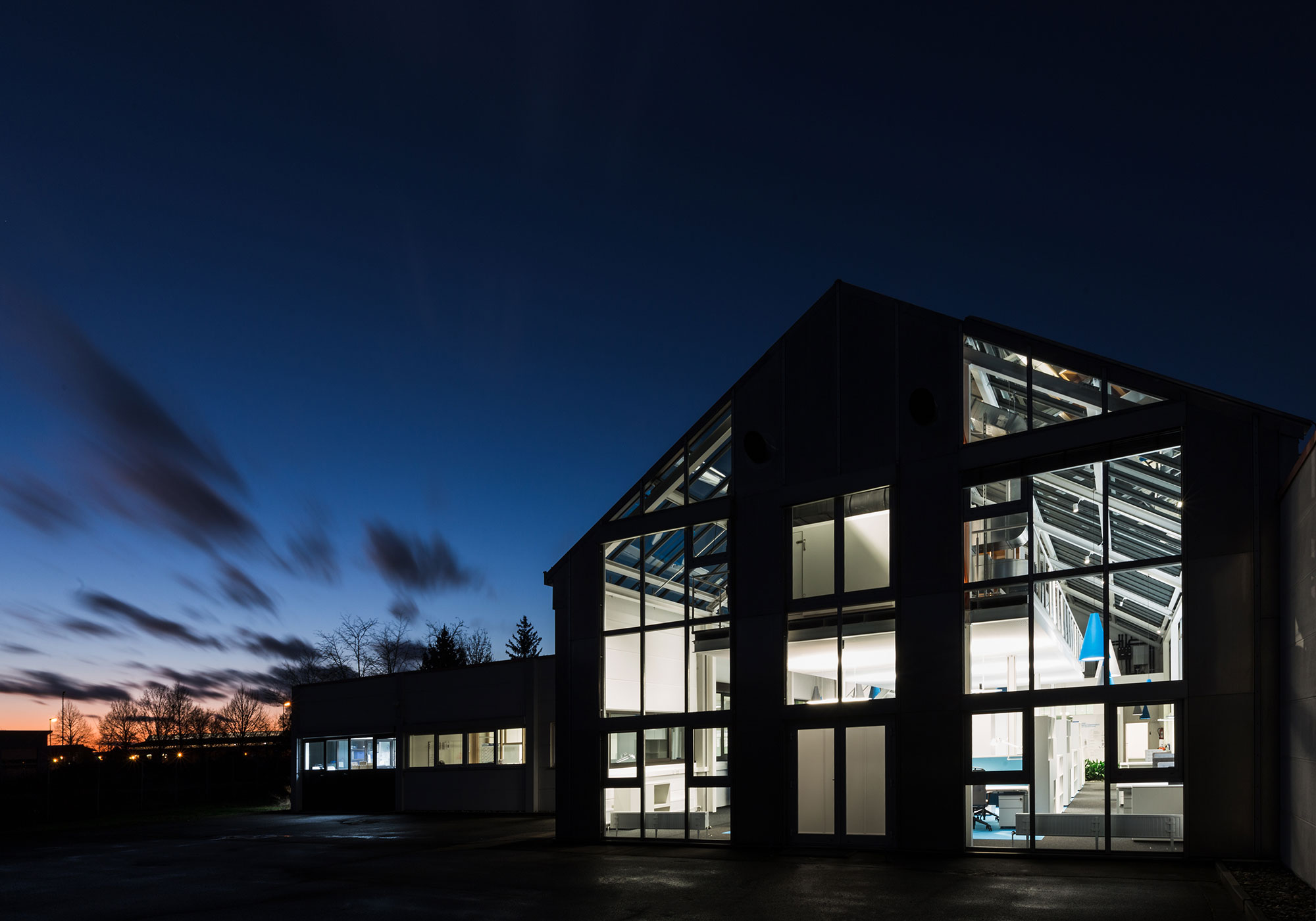 Das moderne BUSSE DESIGN+ENGINEERING Headquarter mit großen Glasfenstern wird von innen beleuchtet und hebt sich vom dunkler werdenden Himmel während der Dämmerung ab. Das Bild zeigt architektonisches Design und natürliche Beleuchtungseffekte.